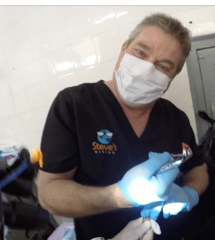 Steve Bender in a mask and Steve's Vision scrubs holds up dental forceps after an extraction