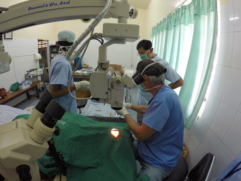 A surgeon operates through a surgical microscope while two masked assistants look on in a tiled operating room