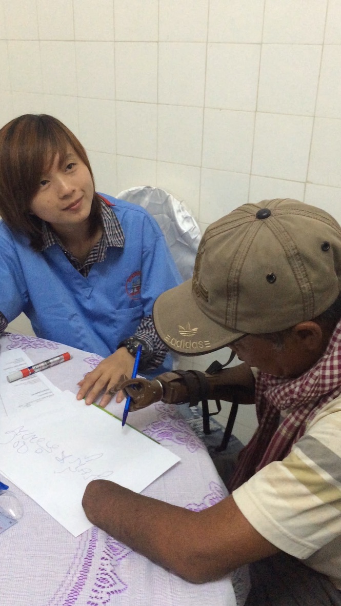 A man wearing a new LN4 prosthetic hand holds a pen and writes a note while a volunteer in a blue uniform watches