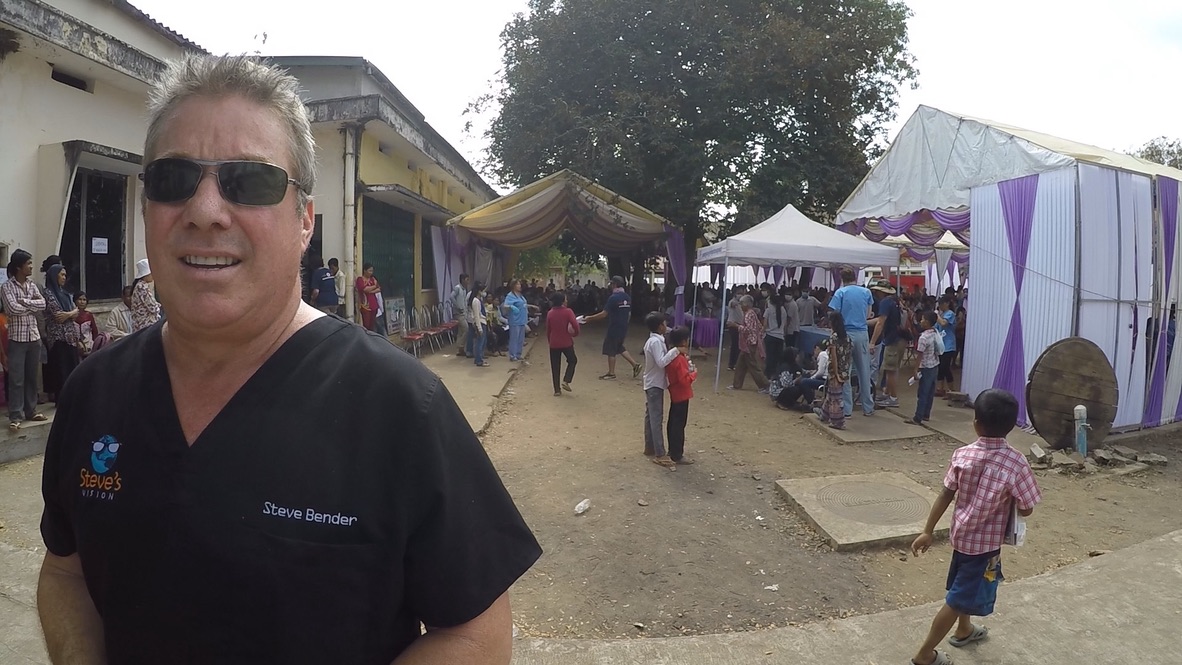 Steve Bender in black scrubs on the clinic grounds, with tents, volunteers and patients moving between stations behind him